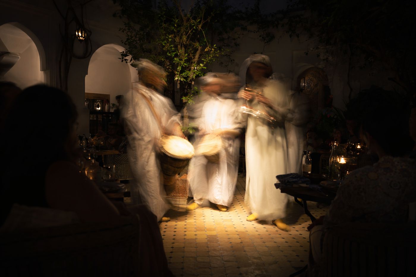 Musiciens flous de mouvement jouant entre les tables d'un patio le soir, silhouettes de convives dans l'ombre