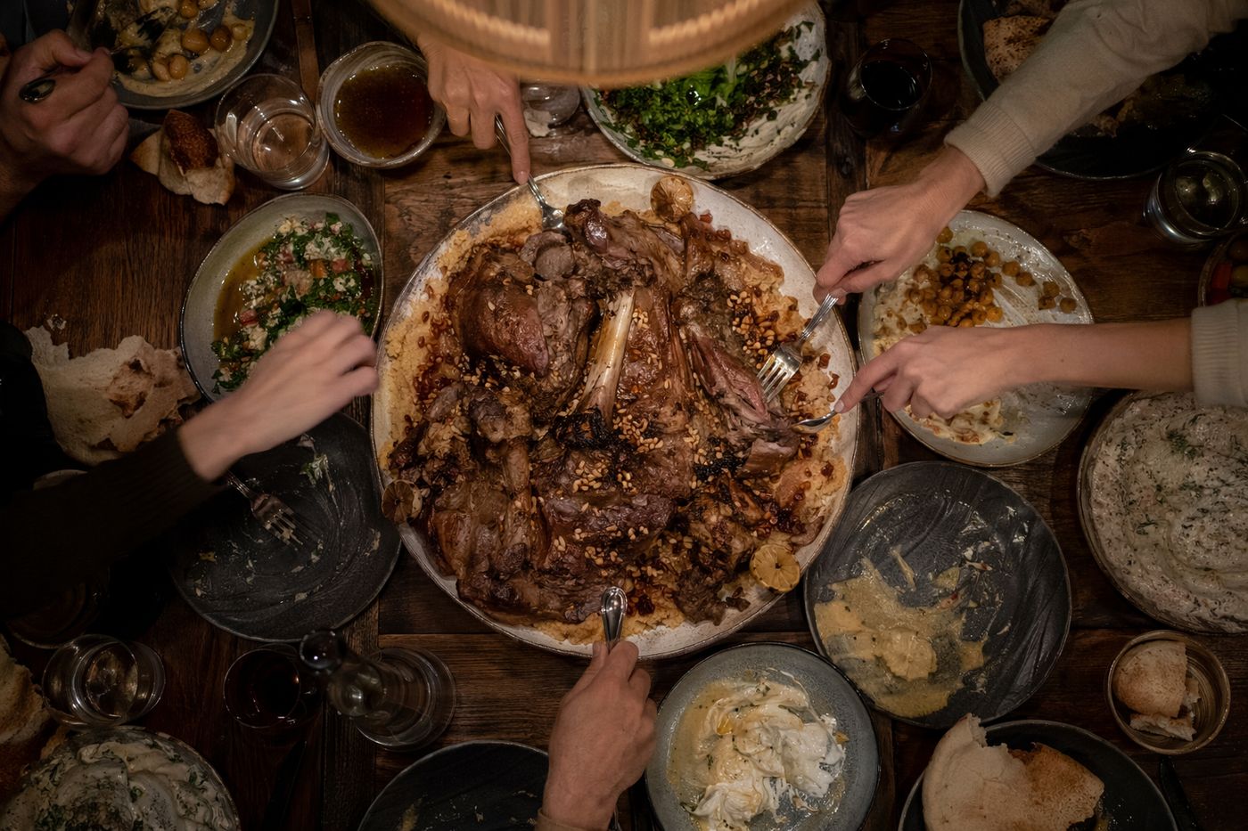 Vue de dessus d'un grand plat d'agneau rôti au centre d'une tablée, plusieurs mains se servant en même temps