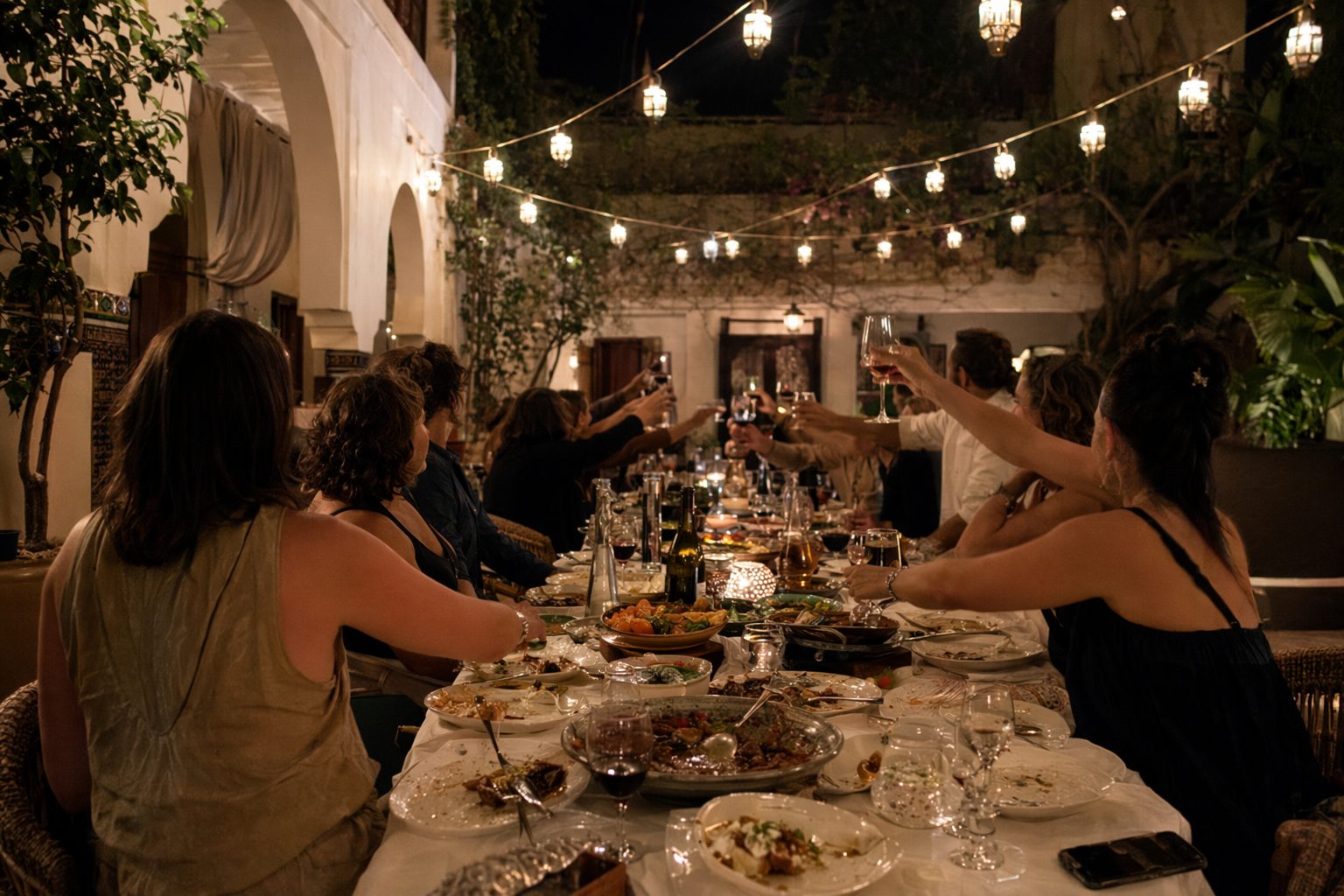 Longue tablée de fête dans un patio le soir, plats à partager au centre et convives de dos sous des lanternes