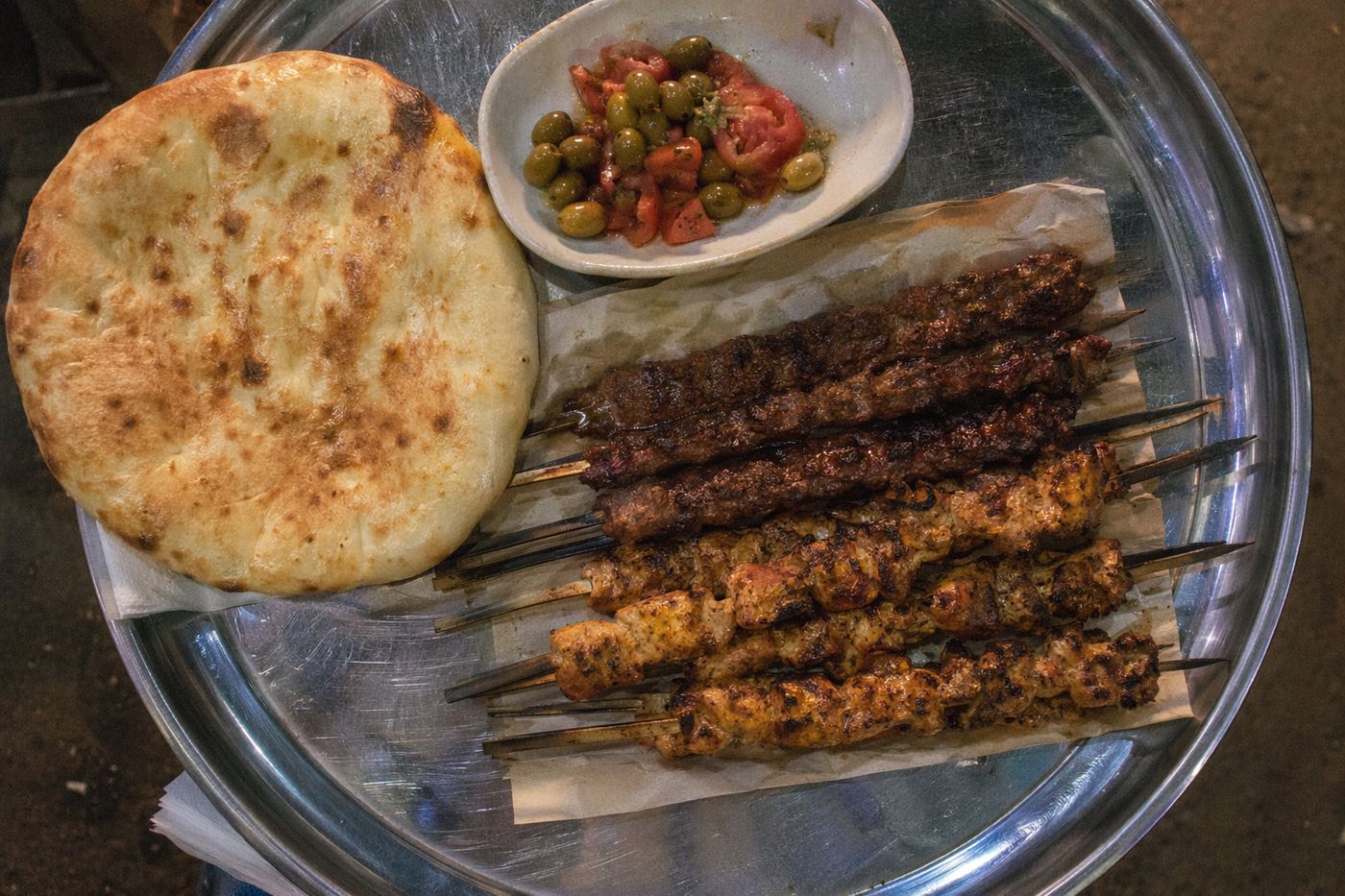 Plateau de brochettes grillées, pain et olives sur un stand de rue éclairé au néon, le soir.