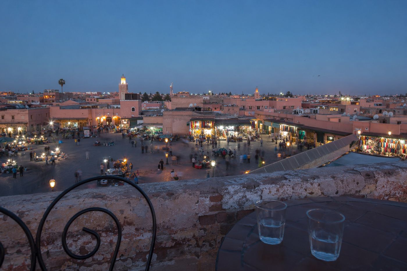 Vue plongeante depuis un parapet de rooftop sur une place animée à l'heure bleue.