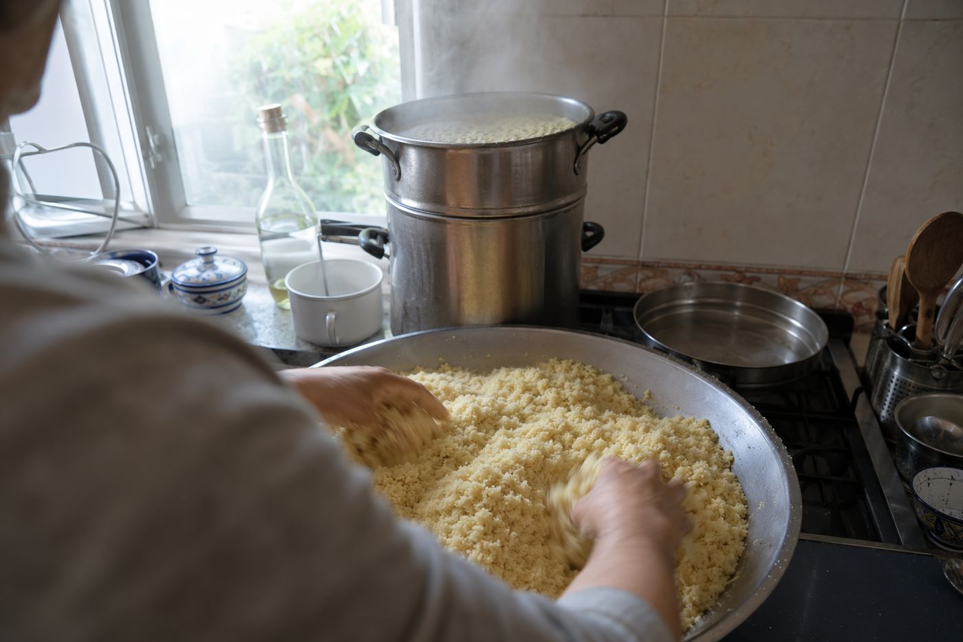 Mains aérant la semoule vapeur dans une grande bassine, couscoussier fumant sur le feu derrière.
