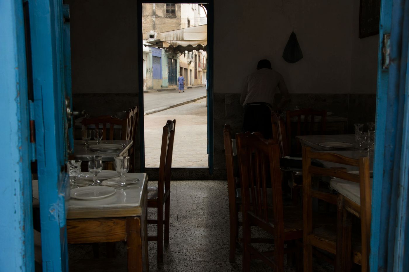 Salle de restaurant sombre en cours de dressage pour le déjeuner, porte ouverte sur la rue lumineuse.
