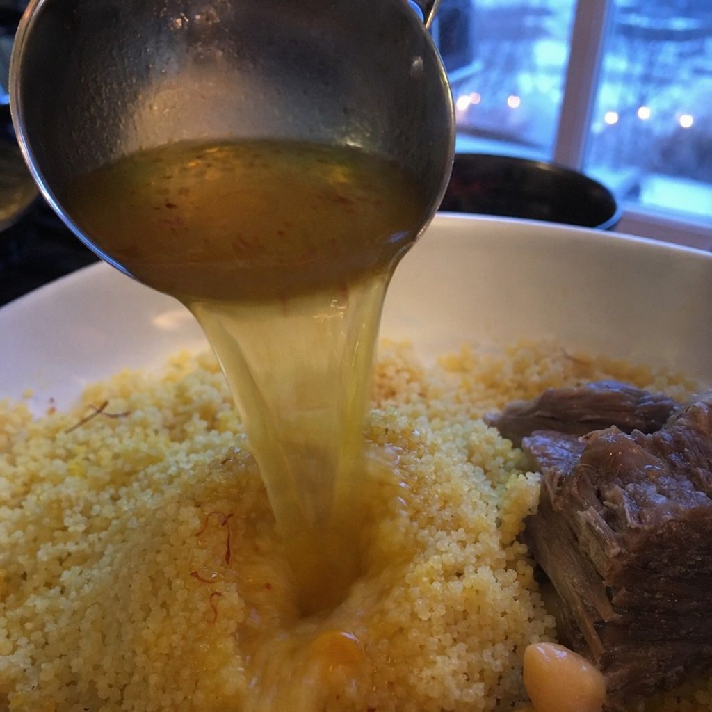 Louche de bouillon clair versée sur des grains de couscous bien détachés, morceau d'agneau au bord.
