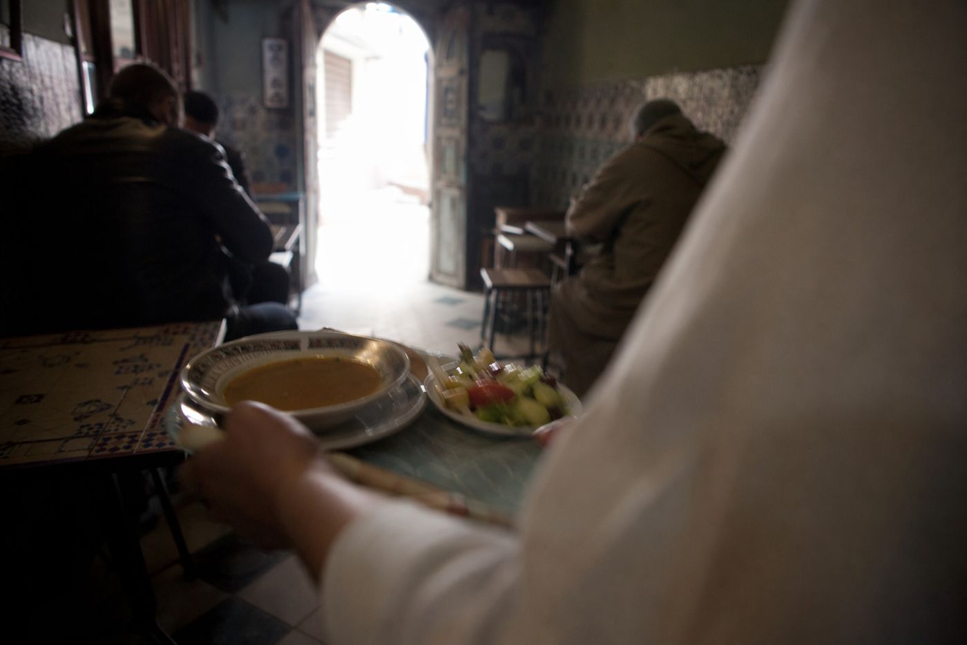 Mains d'un serveur déposant une soupe et une salade sur une table, dans la pénombre d'une petite salle de la médina