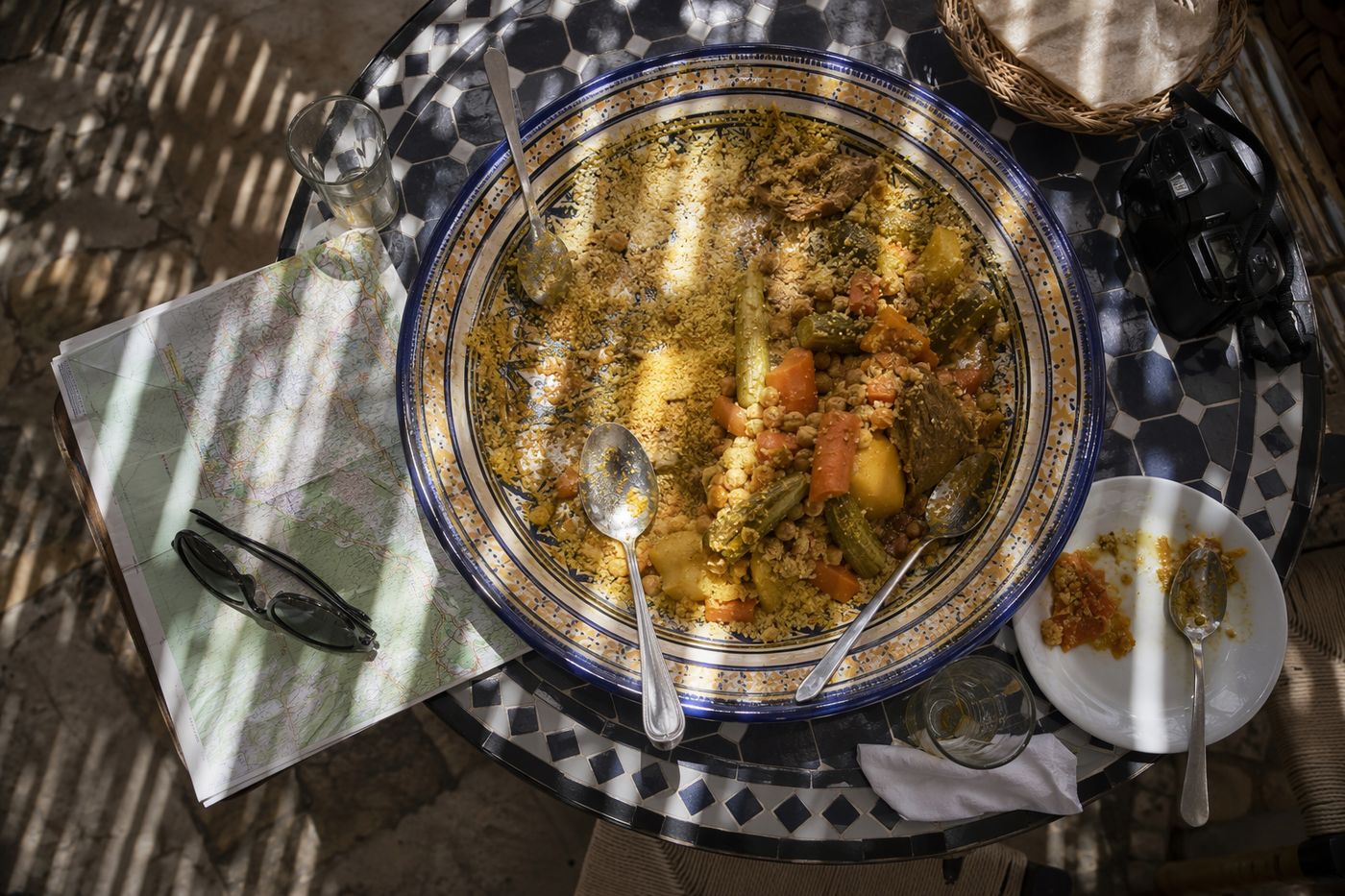 Vue de dessus d'un grand plat de couscous à peine entamé sur une petite table, carte pliée et appareil photo à côté