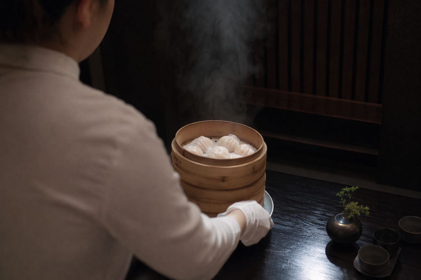 Mains d'un serveur posant des paniers vapeur en bambou sur une table sombre, vapeur montant des dim sum