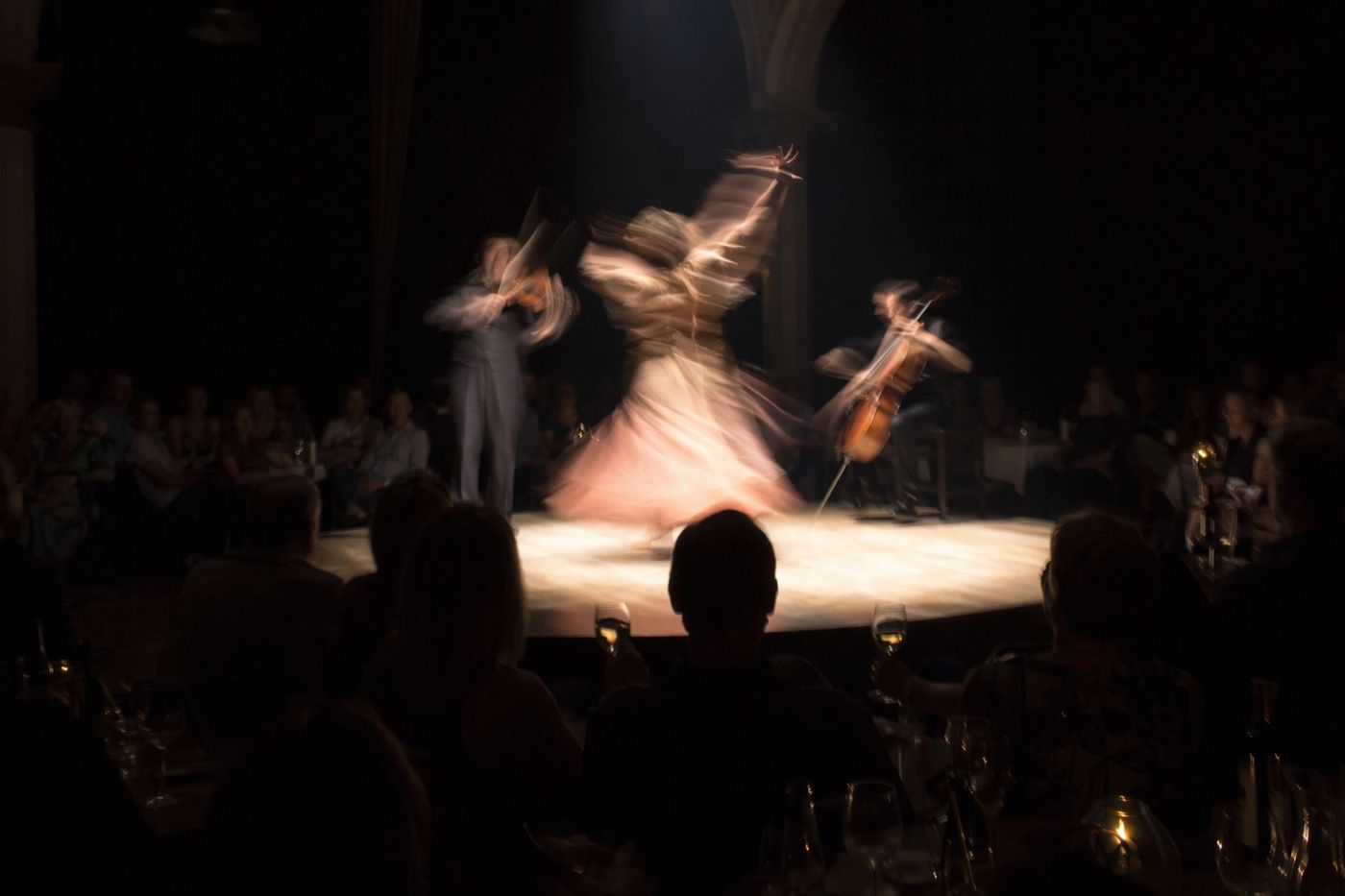 Danseuse et musiciens flous de mouvement sous un projecteur, silhouettes de spectateurs attablés dans l'ombre