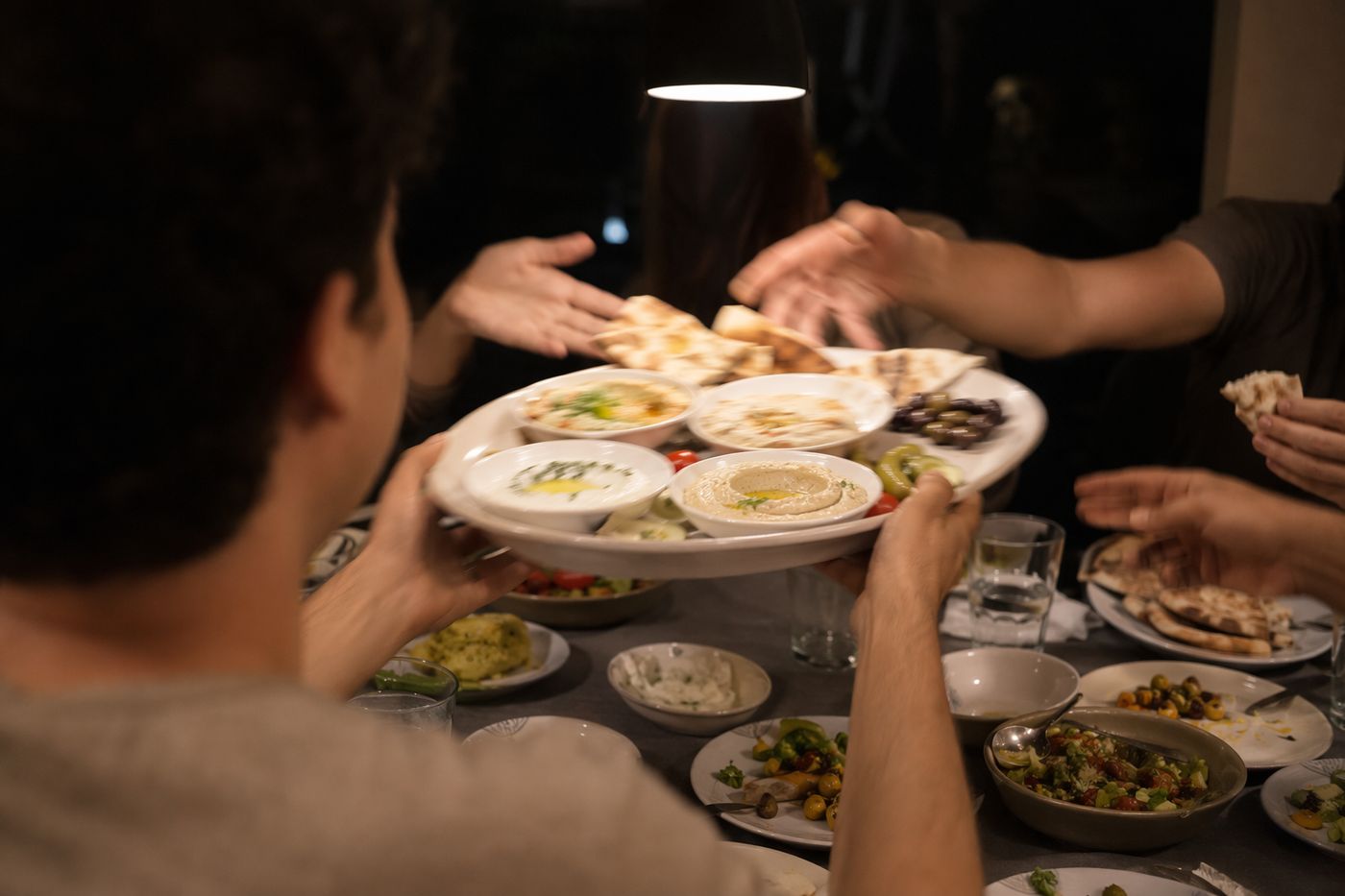 Plateau de mezze passé de main en main au-dessus d'une table le soir, houmous et pain rompu en mouvement