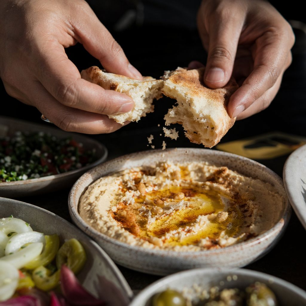 Mains rompant un pain rond au-dessus d'un bol de houmous à l'huile d'olive, miettes en suspension