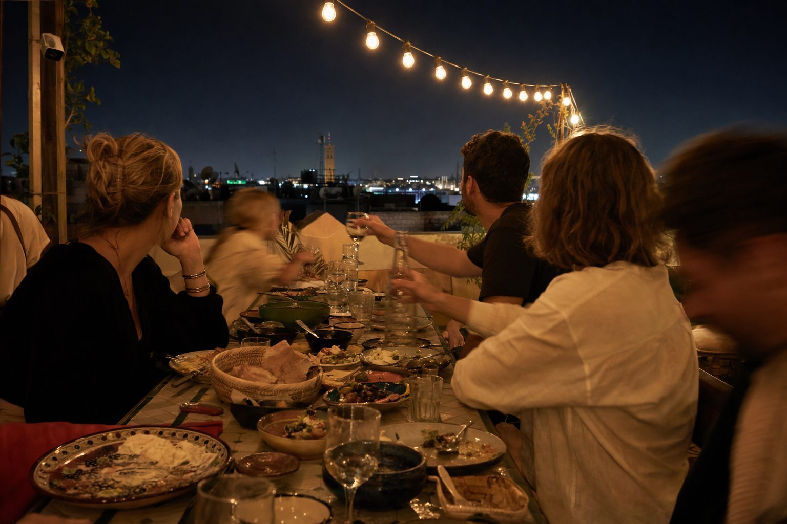 Tablée d'amis de dos sur un rooftop le soir, petites assiettes à partager au centre sous une guirlande d'ampoules