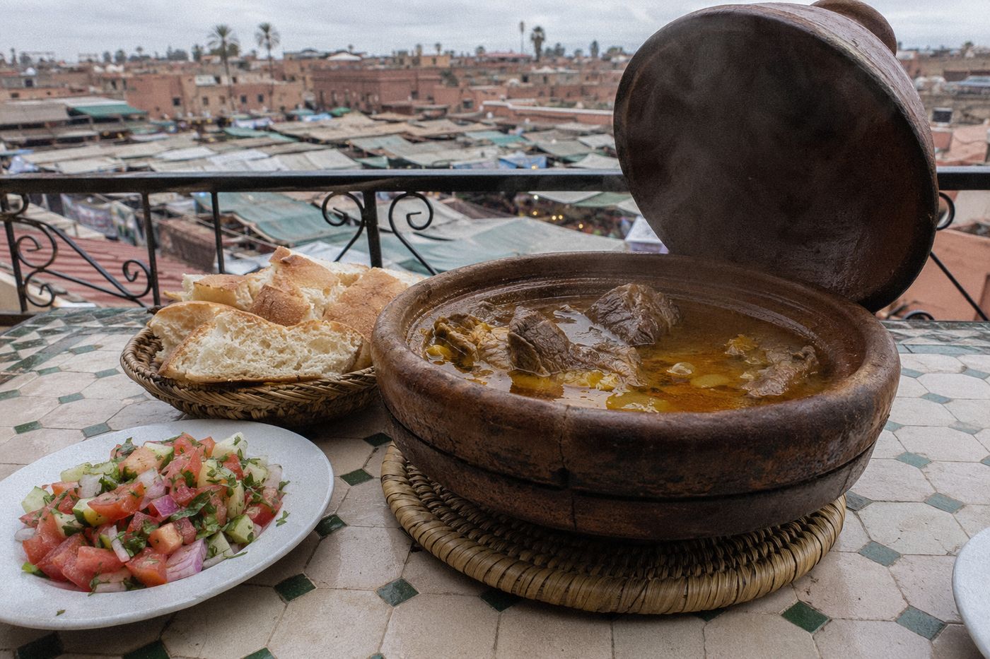 Jarre de tanjia ouverte et fumante sur une table en terrasse, étals d'épices visibles en contrebas.