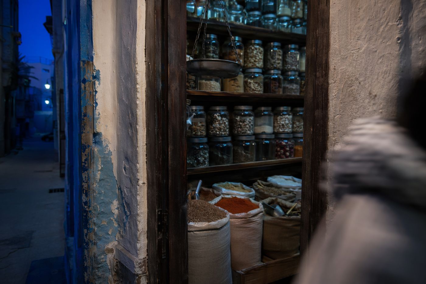 Boutique d'épices vue depuis la porte au crépuscule, bocaux en verre, sacs ouverts et balance suspendue.