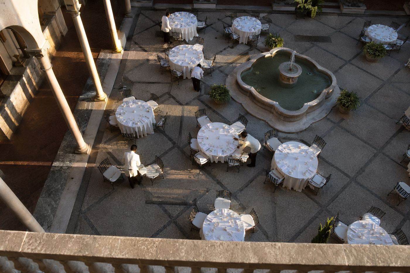 Patio d'un palais-restaurant vu d'en haut depuis une galerie, grandes tables en cours de dressage au soleil rasant