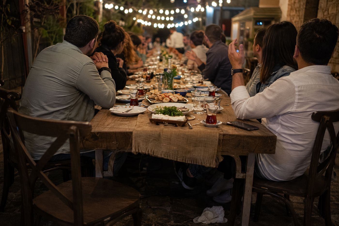 Longue tablée en plein dîner vue depuis un bout de table, plats au centre et convives de dos sous des guirlandes