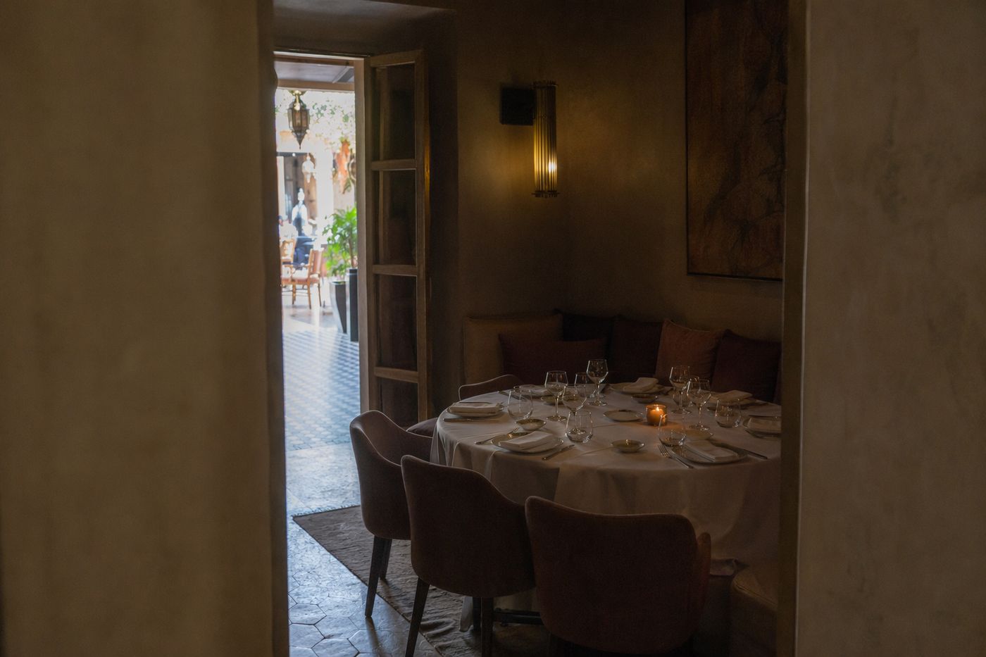 Petit salon calme avec table ronde dressée, vu depuis un couloir, grande salle lumineuse et animée au fond