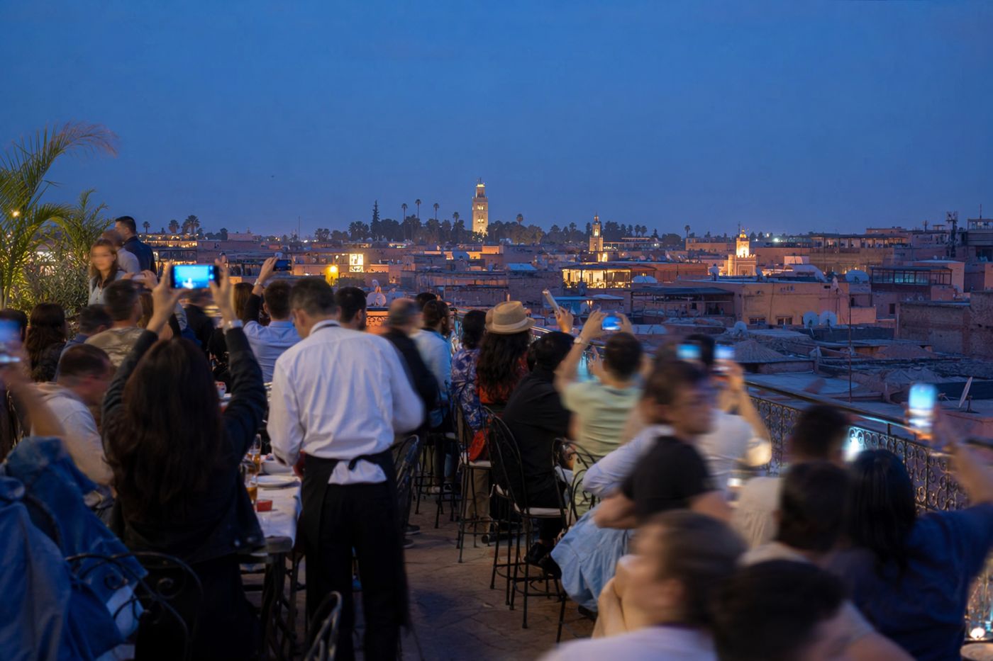 Rooftop bondé à l'heure bleue, tables serrées et silhouettes floues levant leurs téléphones.