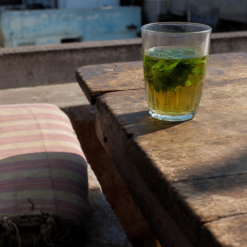Gros plan d'une table en bois usé, coussin rayé et verre de thé sous le soleil dur de midi.