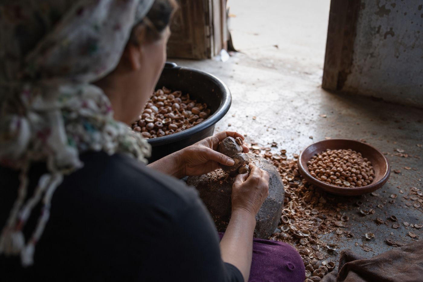 Mains cassant des noix d'argan entre deux pierres, bol d'amandons torréfiés à côté, atelier dans la pénombre.