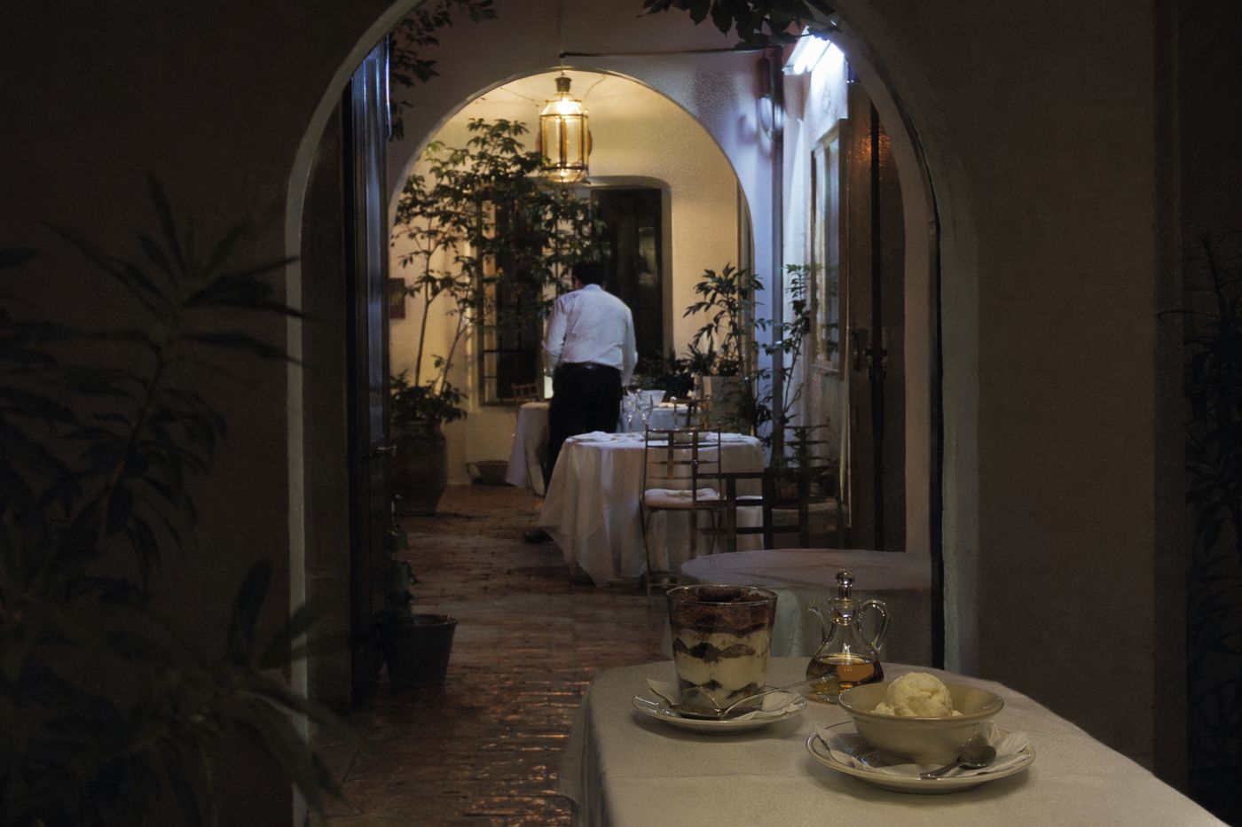 Salle de riad le soir vue d'une arche, dessert au tiramisu et pichet d'huile ambrée posés sur une table.