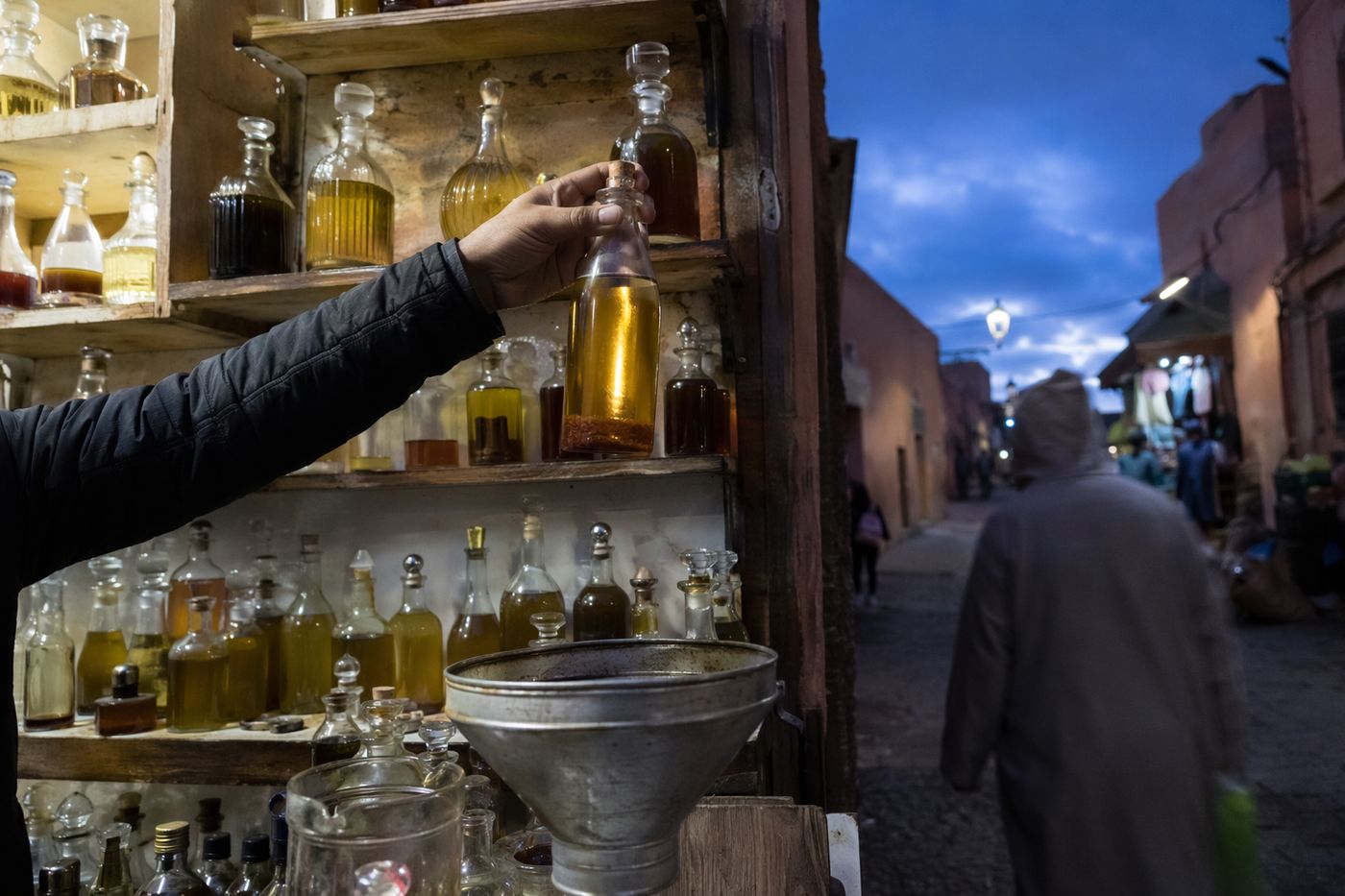 Étal d'huiles au souk au crépuscule, bouteilles ambrées sans étiquette, entonnoir et pichet doseur.