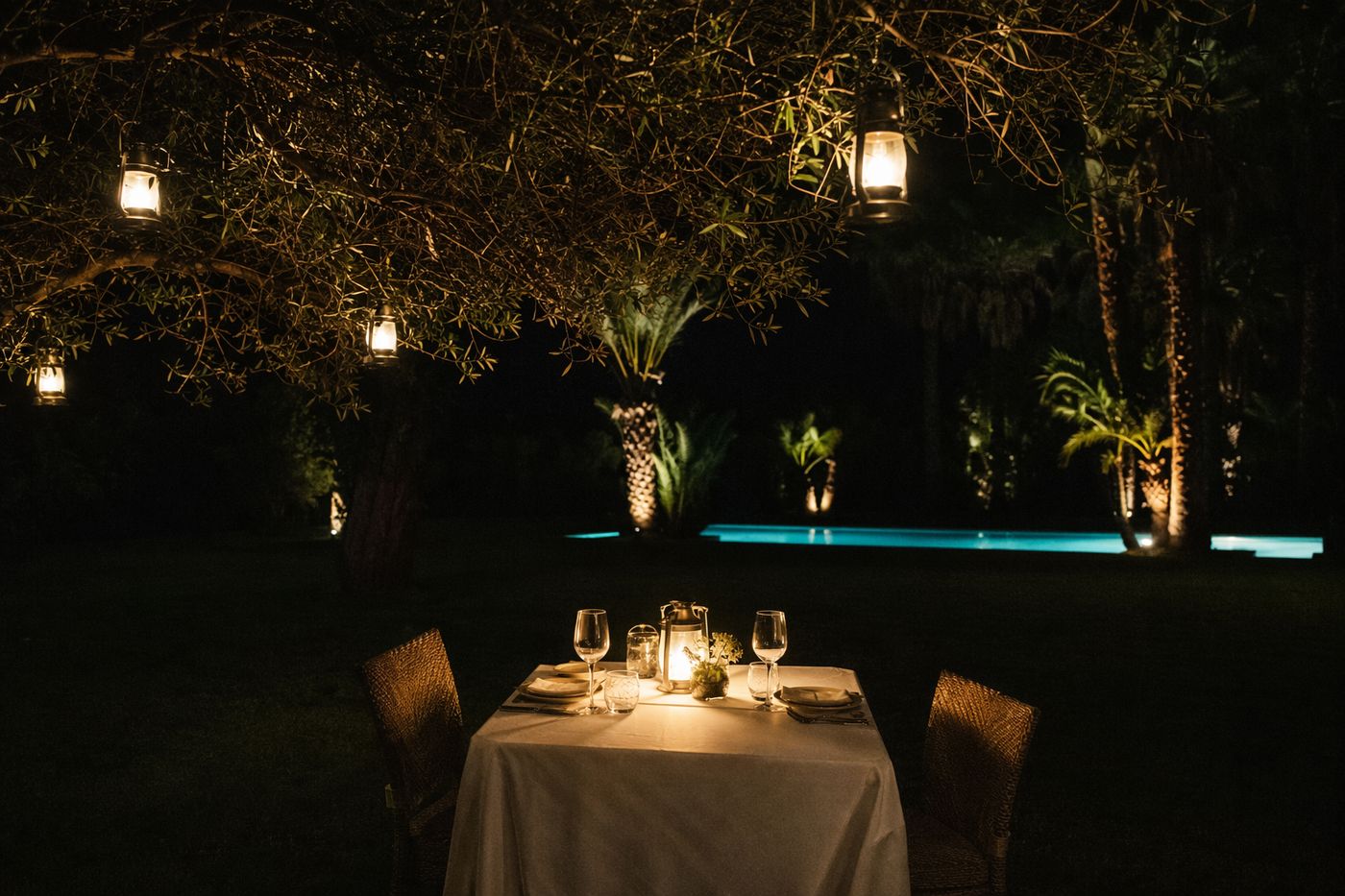 Table dressée le soir dans un jardin, lanternes dans les arbres et piscine éclairée au fond