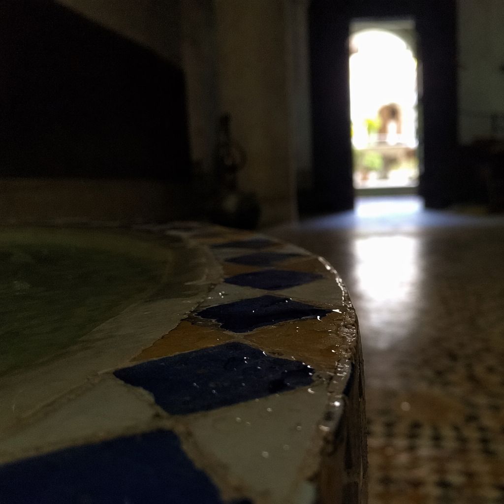 Détail d'une fontaine en zellige dans la pénombre d'un patio de riad, porte lumineuse au fond