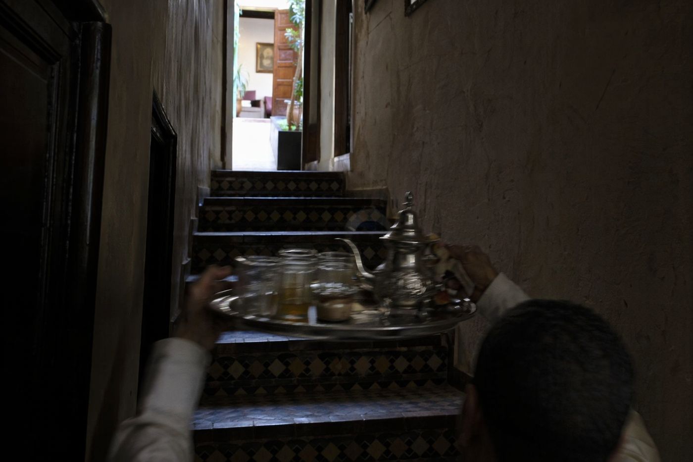 Serveur de dos montant un plateau de thé dans l'escalier sombre d'un riad, porte éclairée en haut