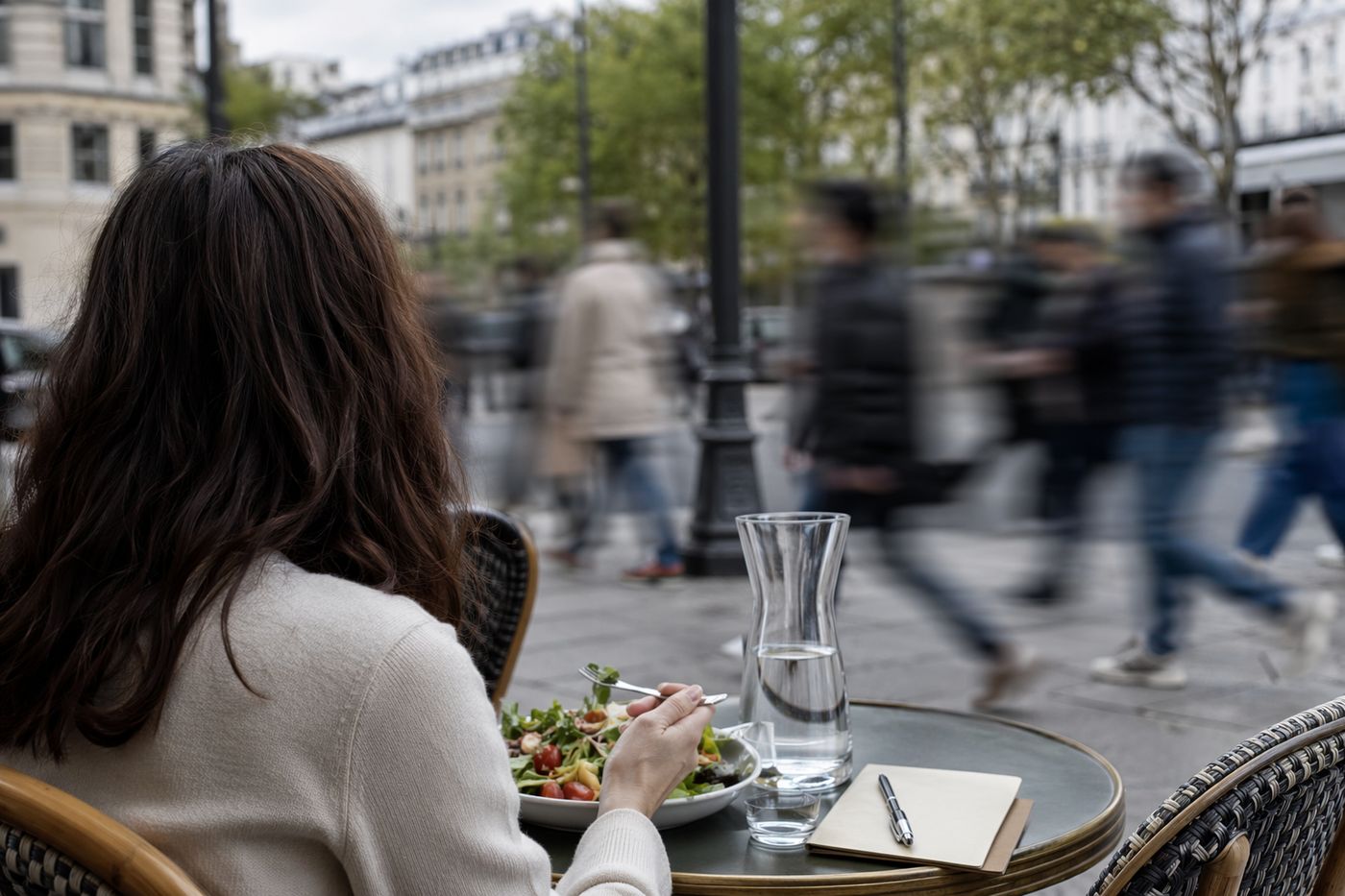 Vue par-dessus l'épaule d'une personne seule en terrasse de trottoir, carnet posé près de l'assiette, rue en mouvement