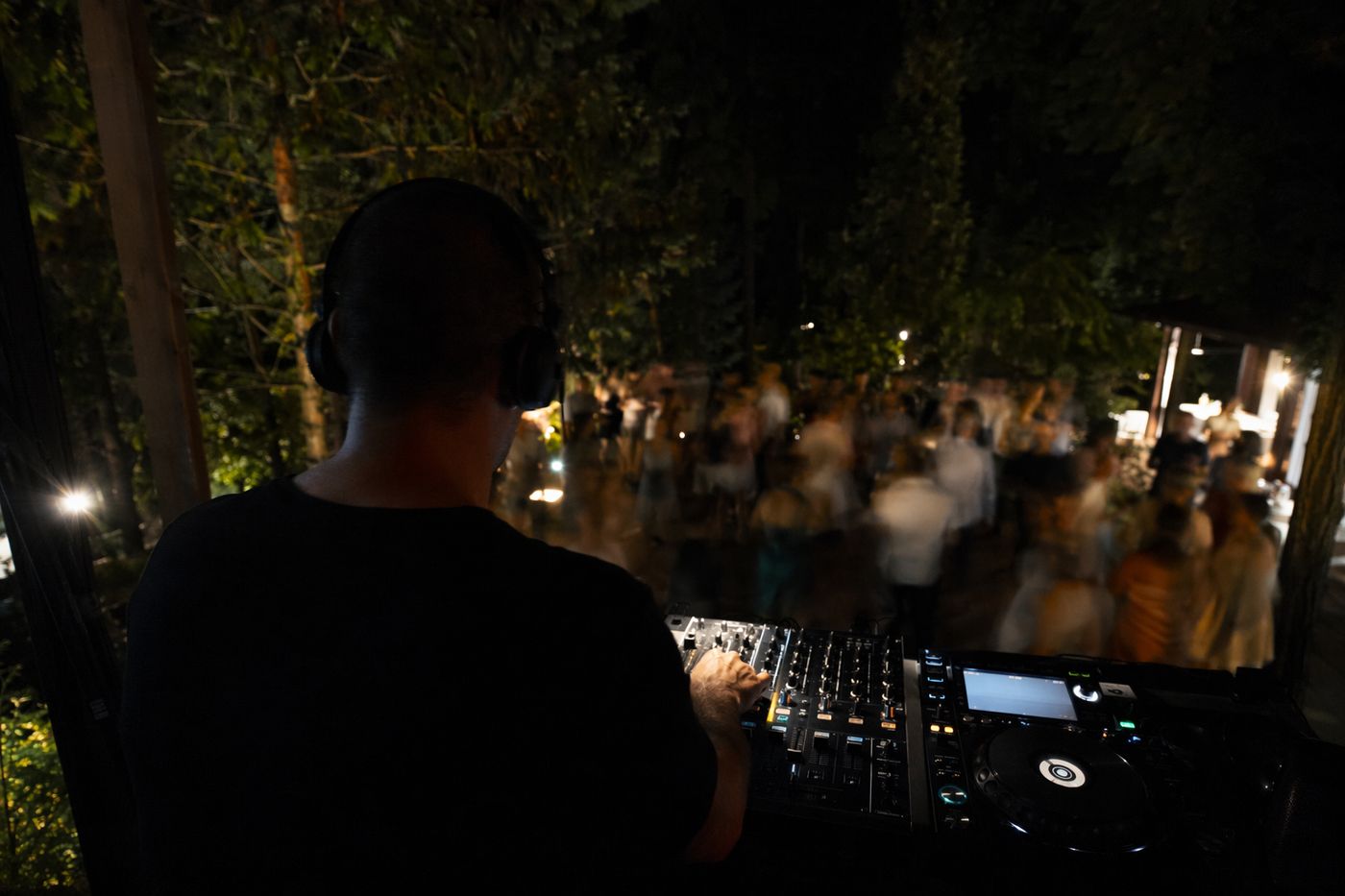 Mains d'un DJ sur une console de nuit, danseurs flous dans un jardin en arrière-plan