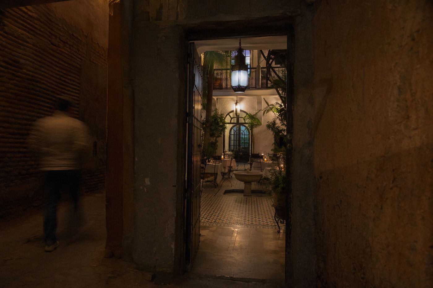 Porte anonyme d'une ruelle sombre ouvrant sur un patio de riad éclairé aux lanternes, le soir.
