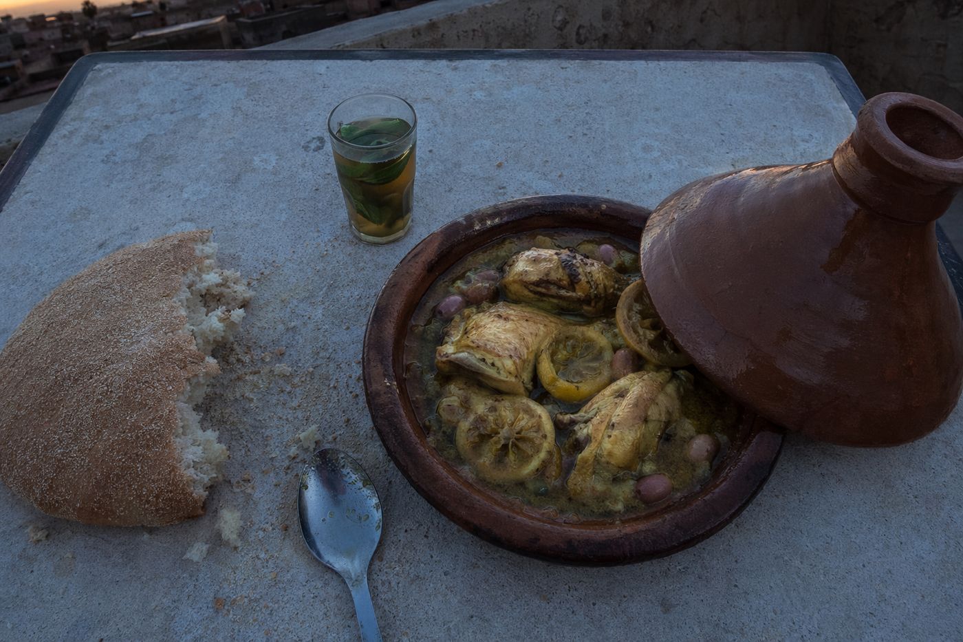 Vue du dessus d'un tajine de poulet au citron, pain et thé sur une table simple de rooftop au crépuscule.