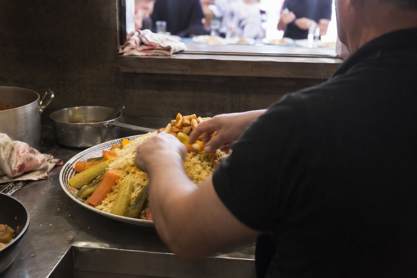 Mains d'un cuisinier dressant un plat de couscous aux légumes dans une cuisine sombre.