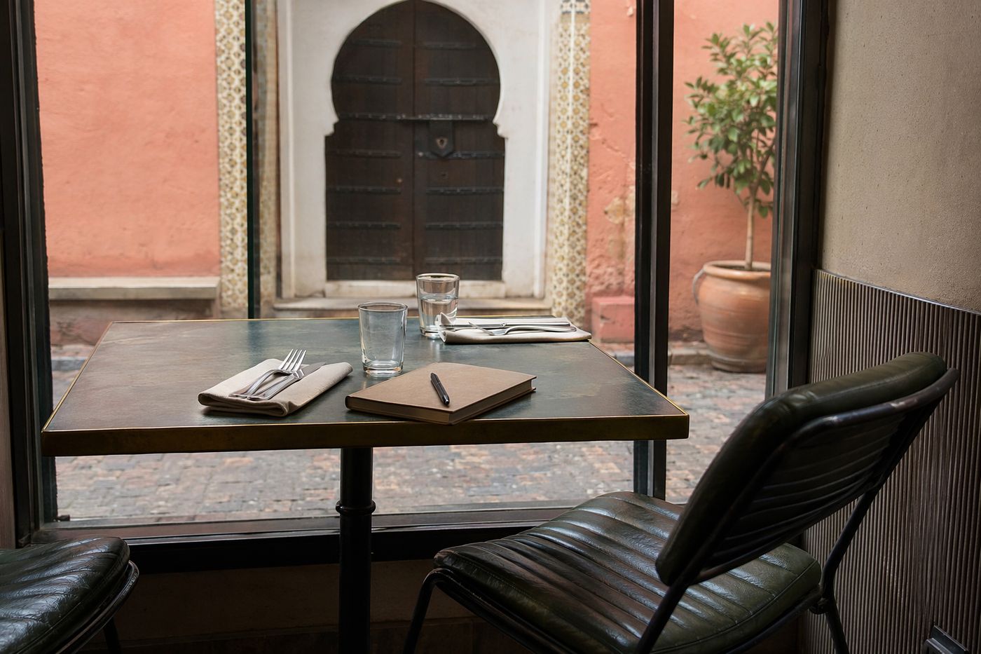 Petite table de restaurant près d'une fenêtre, carnet fermé et stylo posés à côté d'un verre d'eau.
