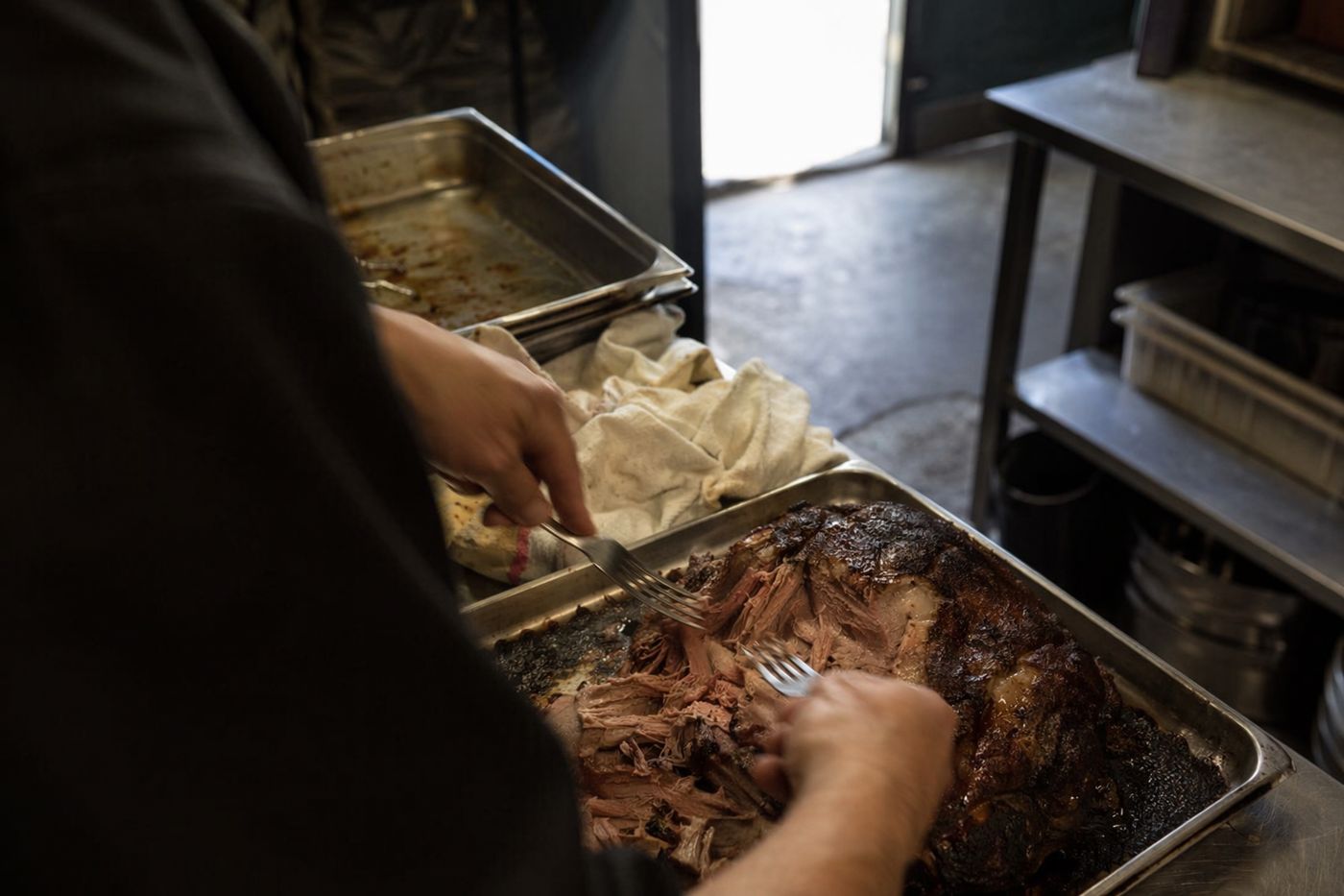 Mains effilochant une épaule d'agneau rôtie lentement sur un plateau, dans une cuisine sombre.