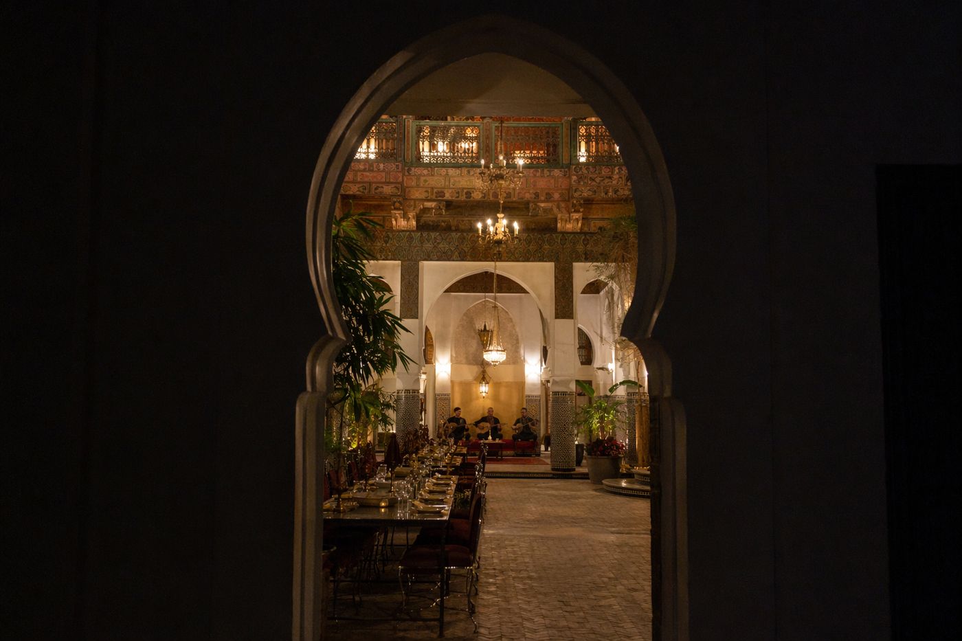 Patio de riad illuminé pour le dîner, musiciens au fond, vu depuis un couloir sombre