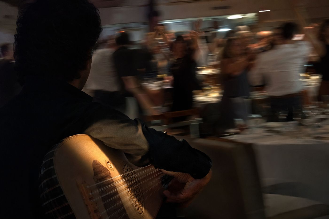 Mains d'un joueur d'oud en gros plan, convives flous dansant entre les tables derrière lui