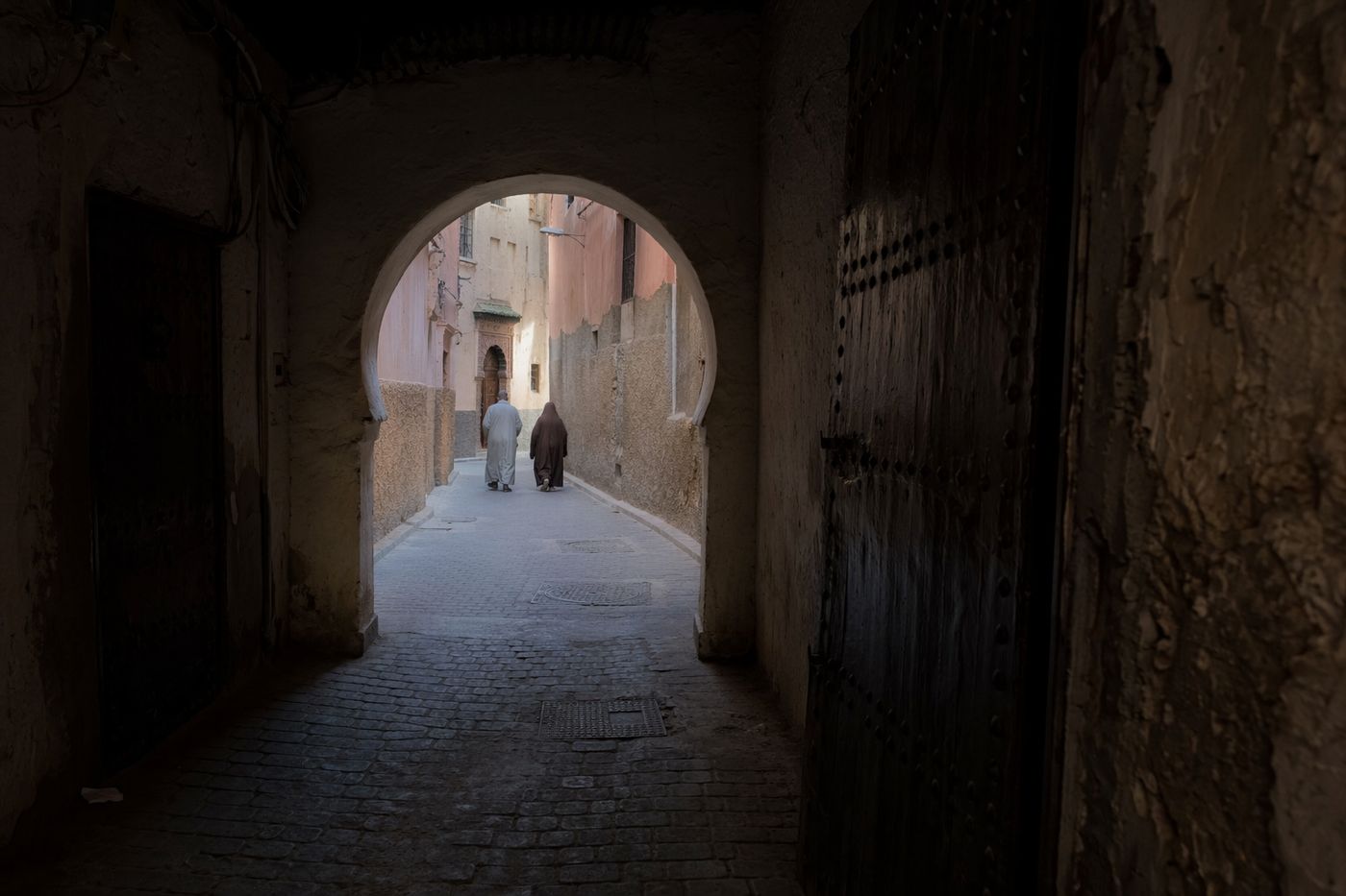 Passage couvert et sombre de la médina ouvrant sur une ruelle ensoleillée, silhouettes de dos