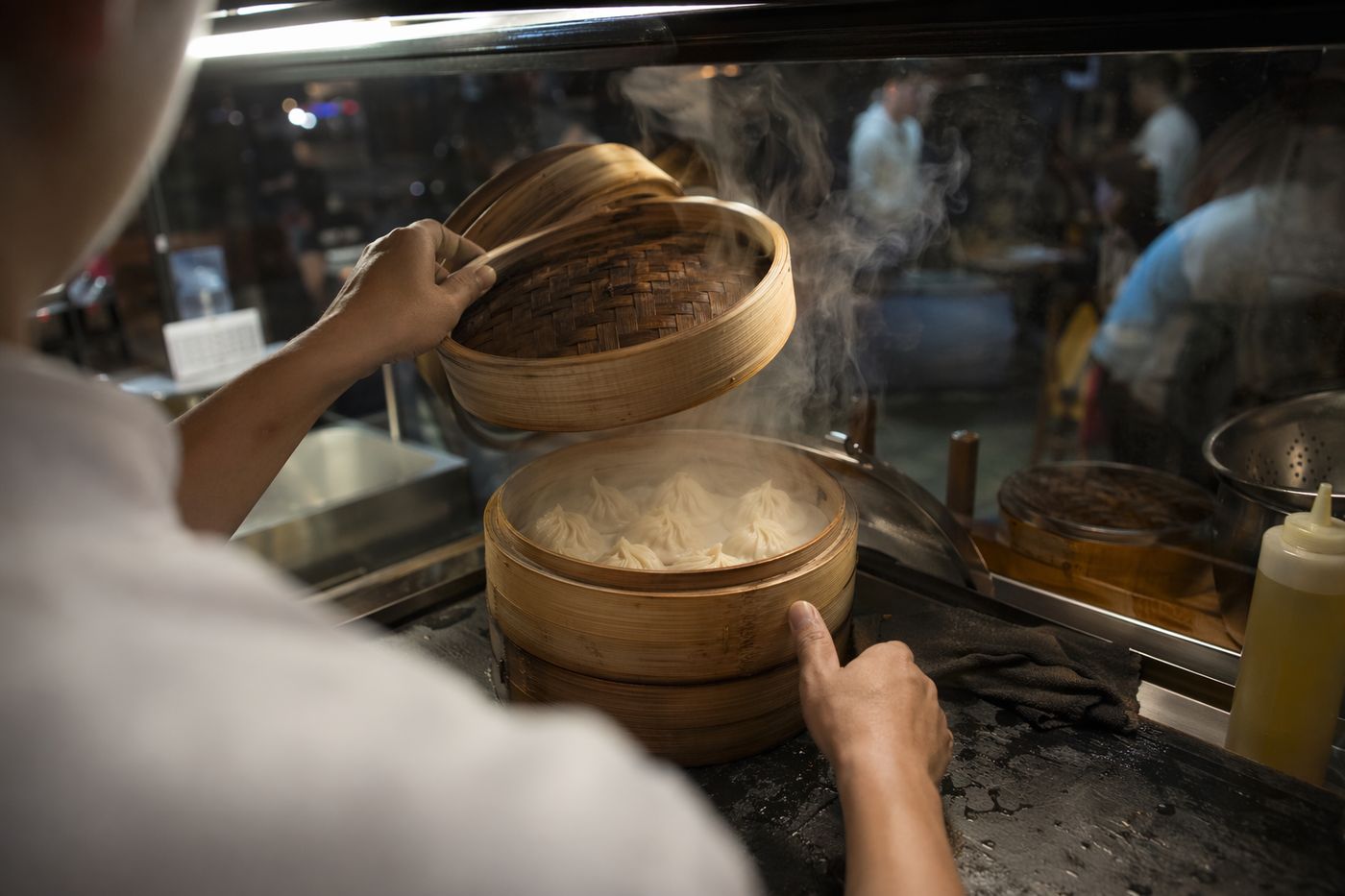 Mains soulevant le couvercle d'un panier vapeur en bambou rempli de dumplings, vapeur visible.