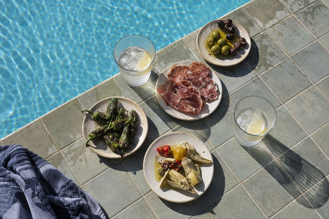 Assiettes de tapas et verres au bord d'une piscine sous le soleil de midi, reflets d'eau sur la table.