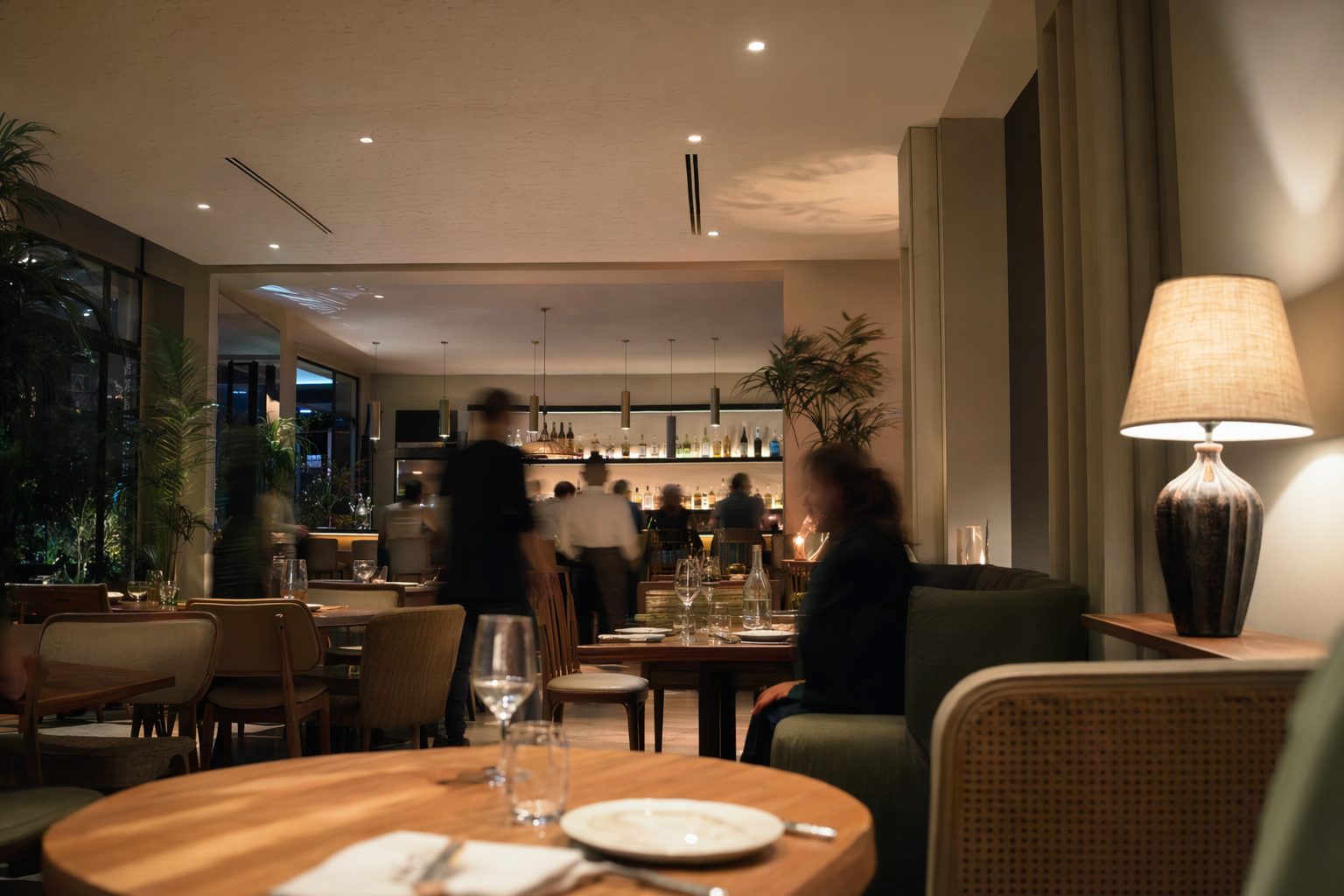 Salle de restaurant contemporaine le soir, mobilier épuré et silhouettes floues entre les tables.
