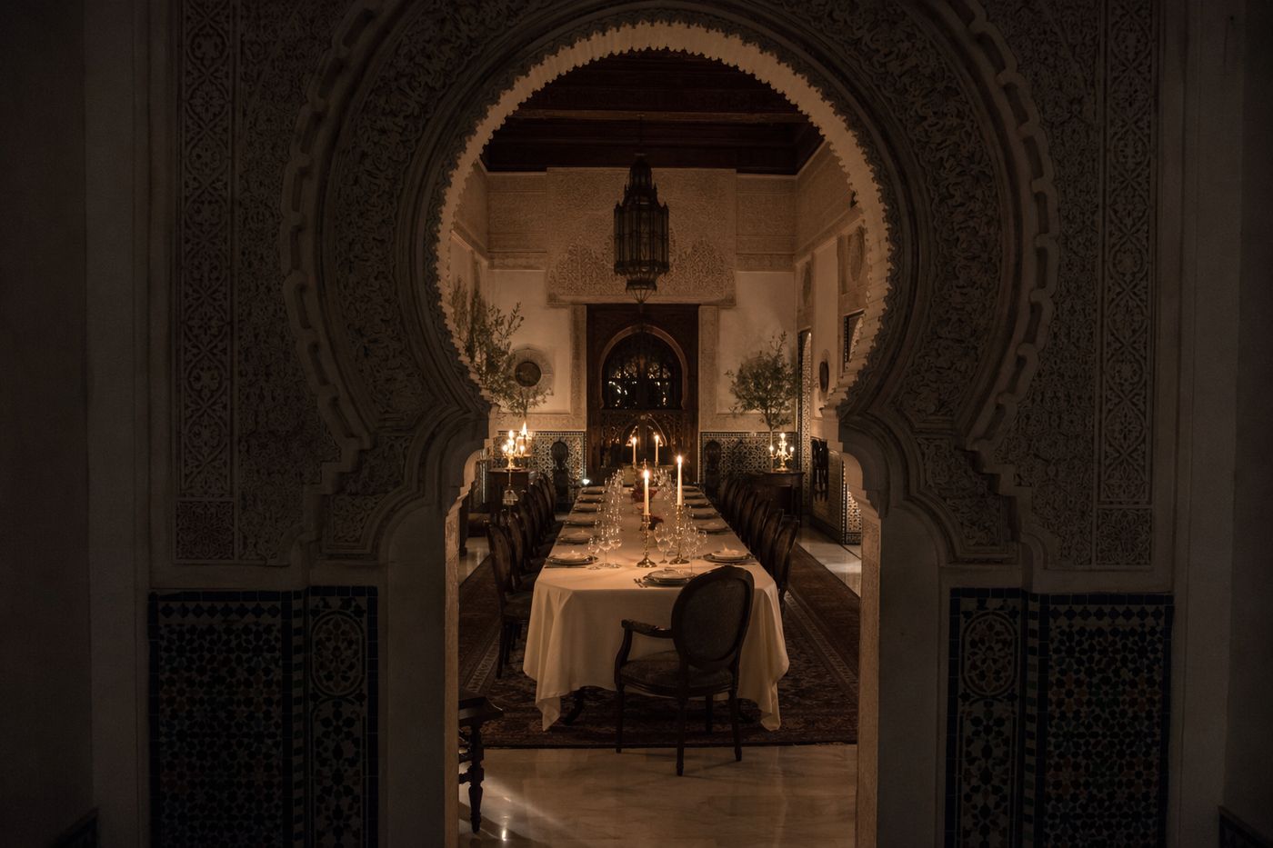 Salon de palais aux bougies vu à travers une arche sculptée, zellige et table nappée de blanc.