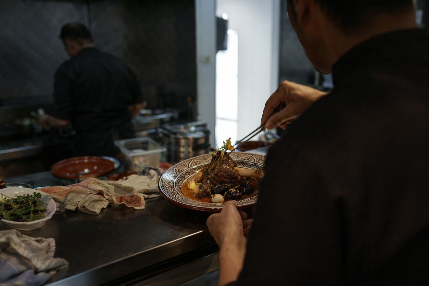 Mains d'un cuisinier dressant une assiette marocaine raffinée au passe d'une cuisine sombre.