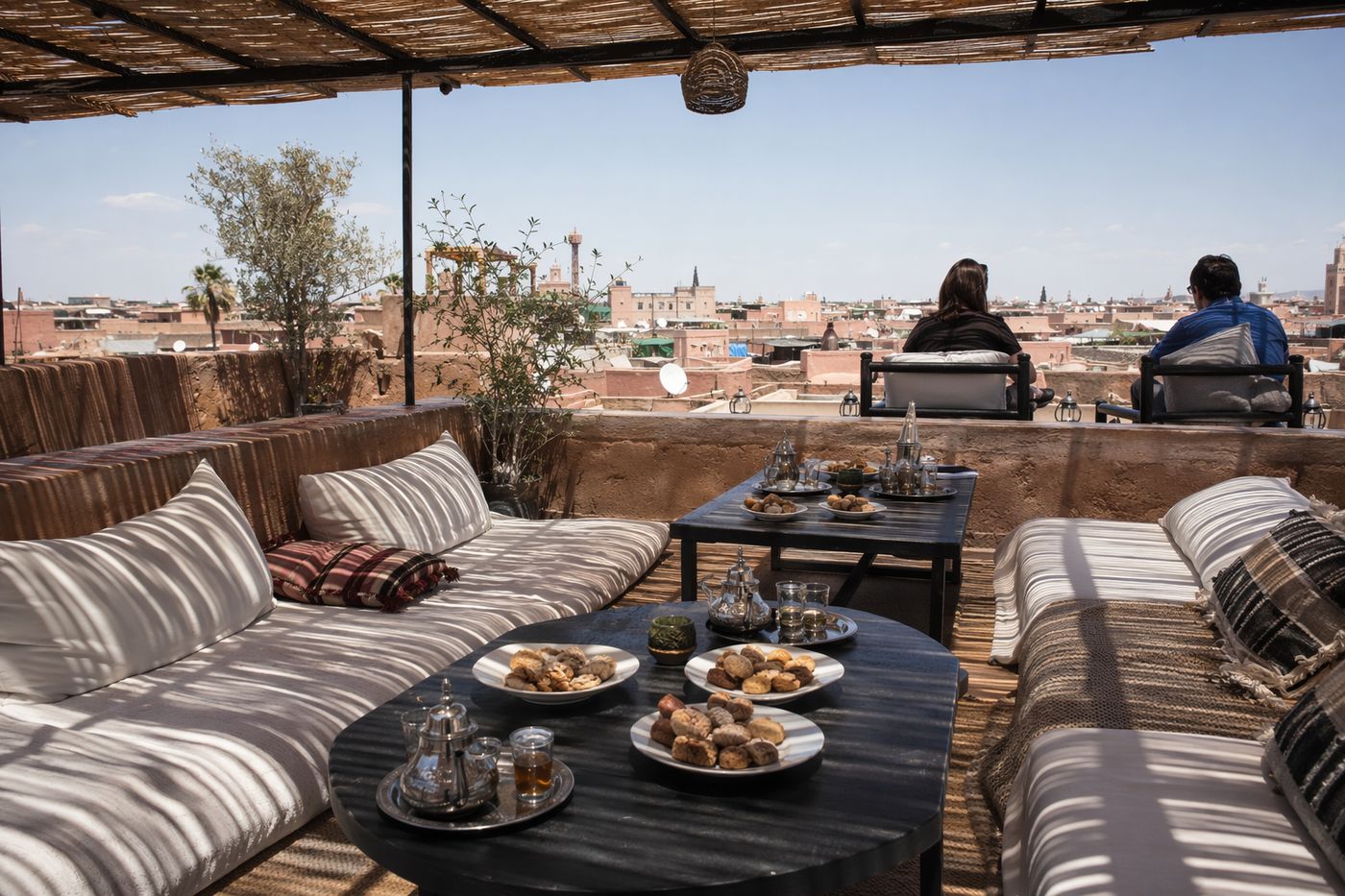 Terrasse de rooftop l'après-midi, assiettes de pâtisseries marocaines et théières sous des claies d'ombrage.