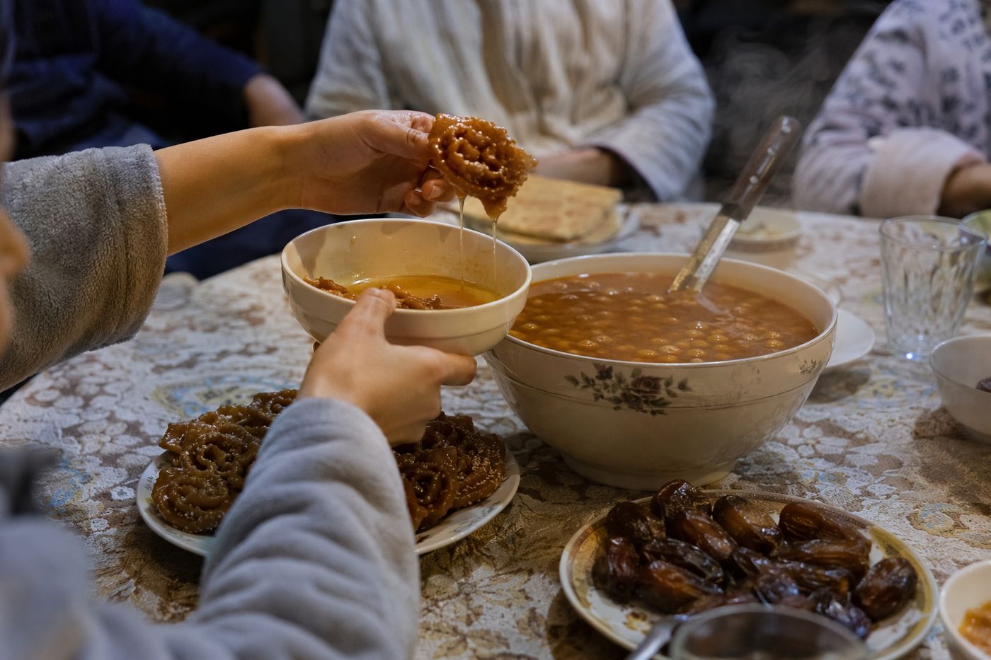 Mains sortant une chebakia du miel chaud près d'un bol de harira fumante et de dattes, table du soir.