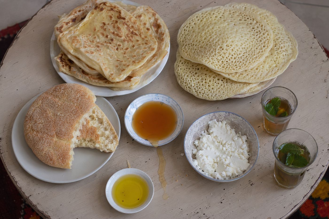 Vue du dessus d'une table ronde de petit-déjeuner marocain : crêpes, batbout ouvert, miel, fromage frais et thé.