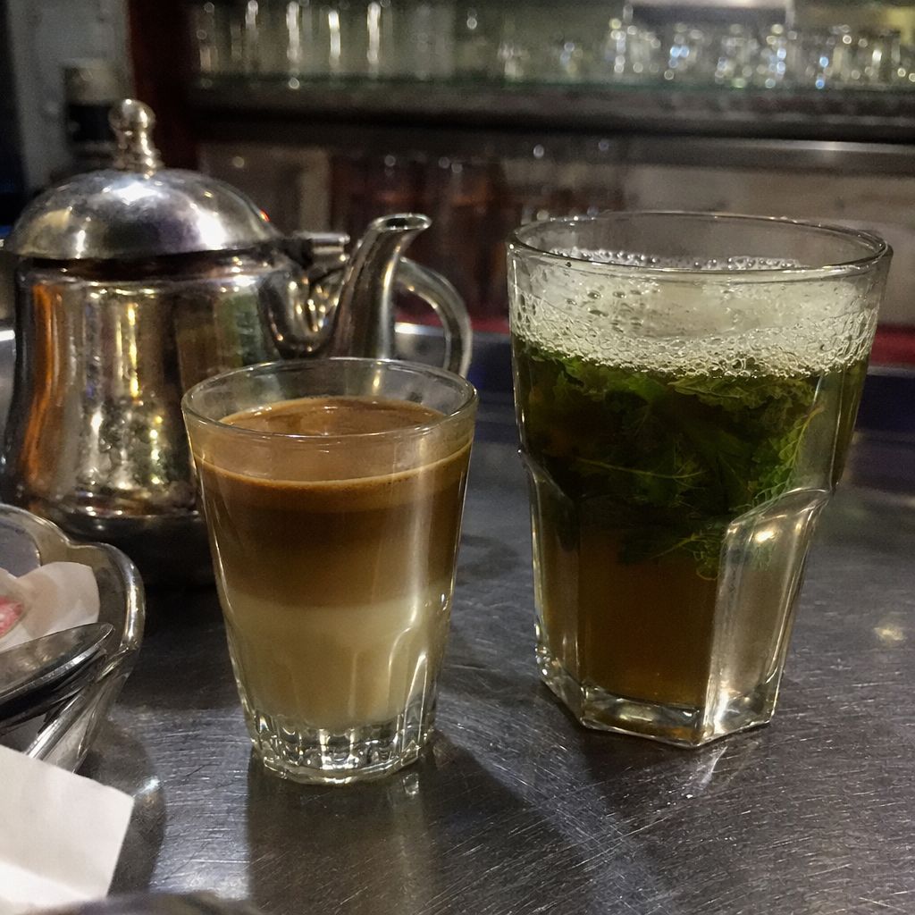 Gros plan sur un verre de nos-nos aux couches lait-café, théière et verre de thé à la menthe sur un comptoir de café.