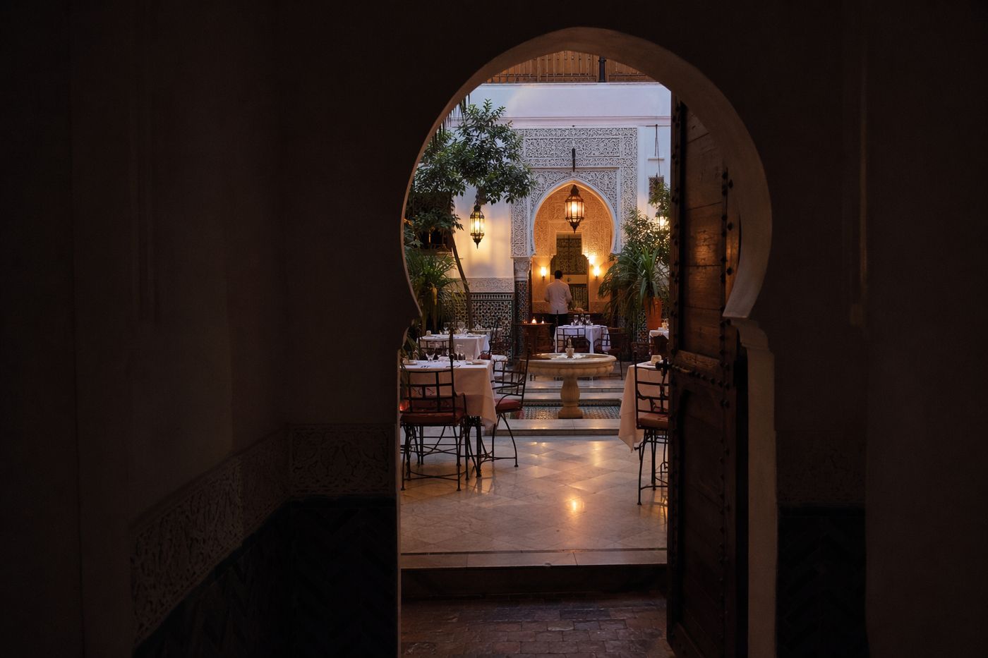 Patio d'un riad-restaurant vu à travers une porte en arc, tables dressées, fontaine et zelliges sous les lanternes