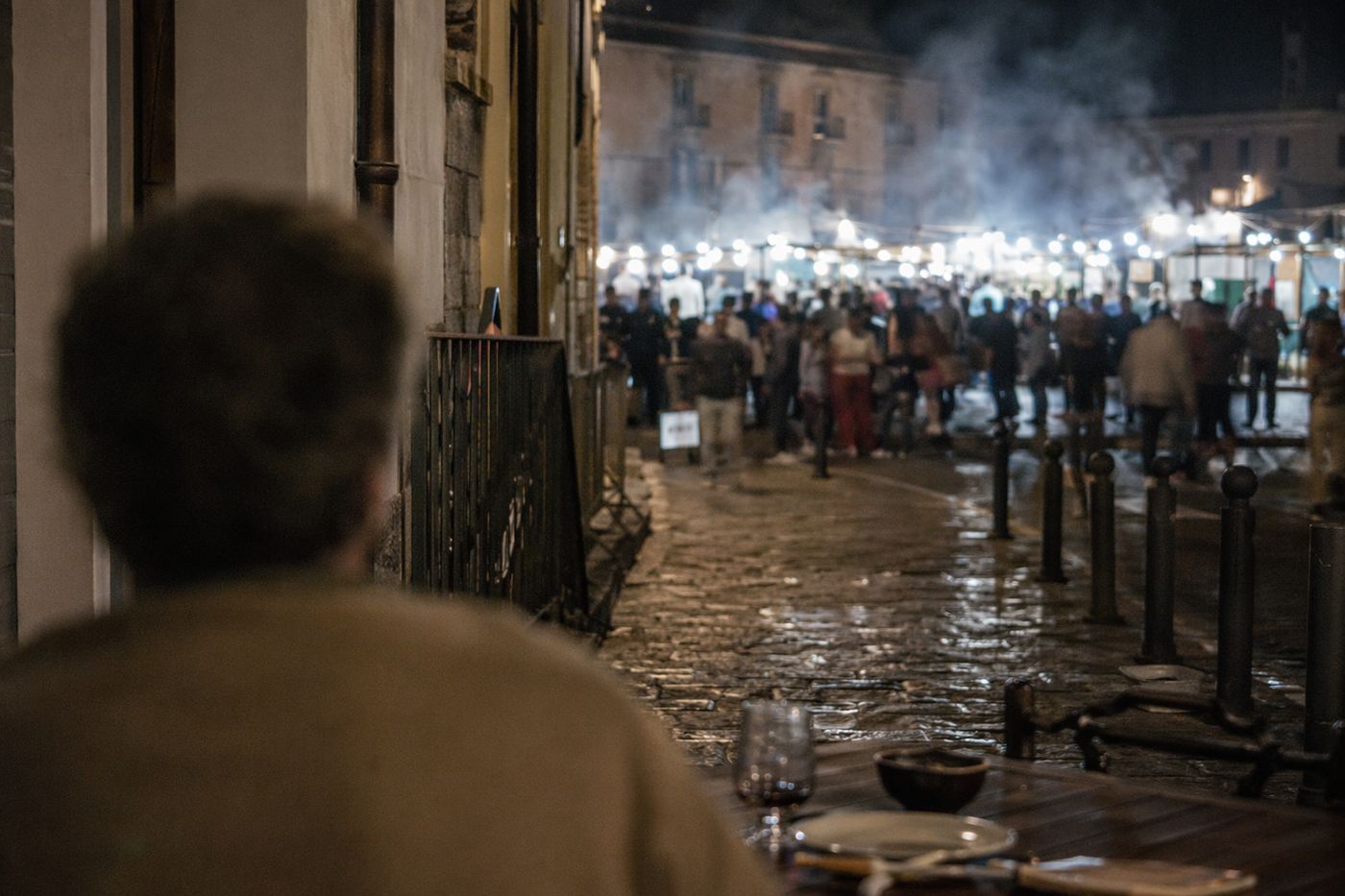 Depuis une table calme en retrait, vue de nuit sur la fumée et les lumières des stands d'une grande place animée