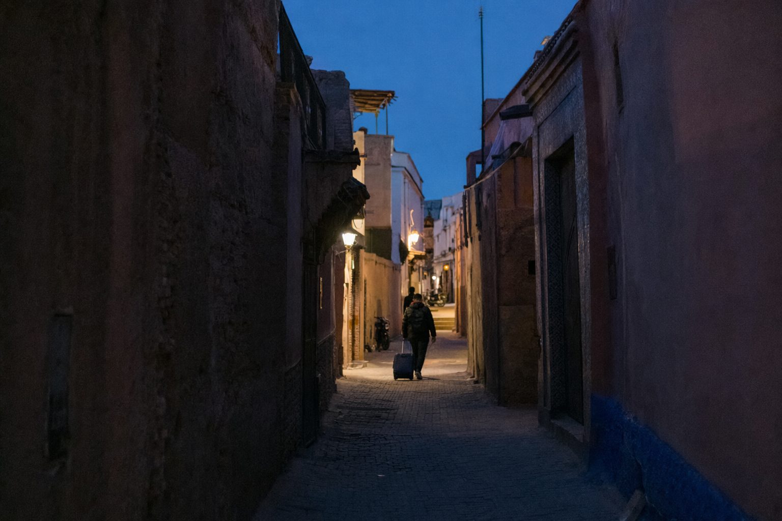 Ruelle de la médina à l'heure bleue, voyageur de dos tirant une valise vers une sortie éclairée